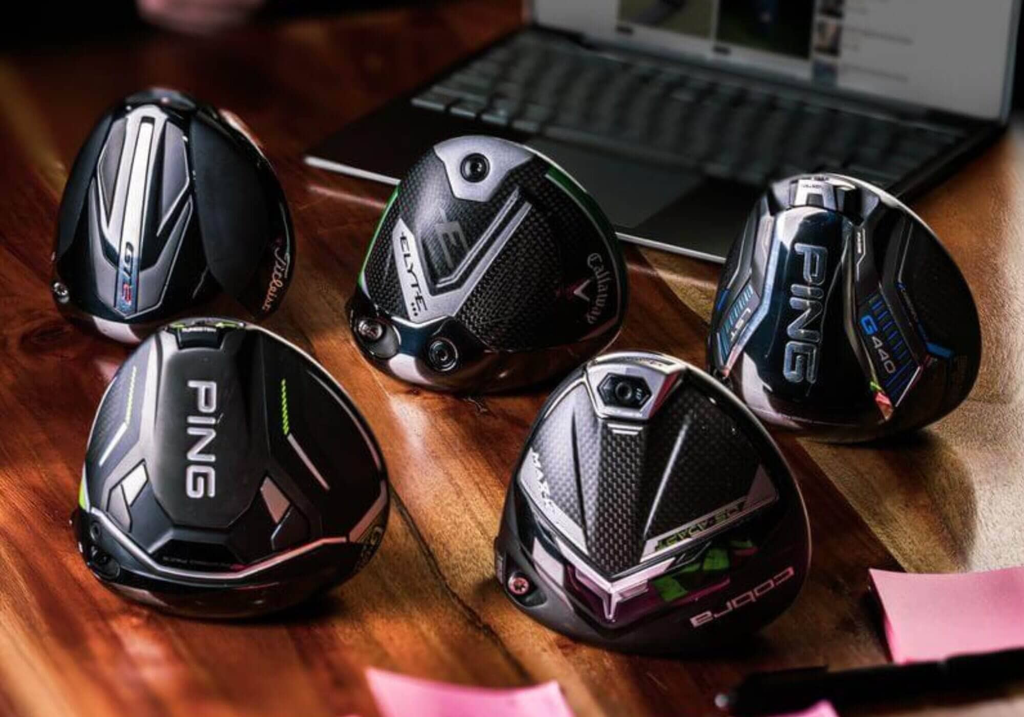 PING、キャロウェイ、タイトリスト、コブラなど、2025年モデルの主要ドライバーヘッドが机上に並ぶ。背景にノートPCとレビュー動画も見える。