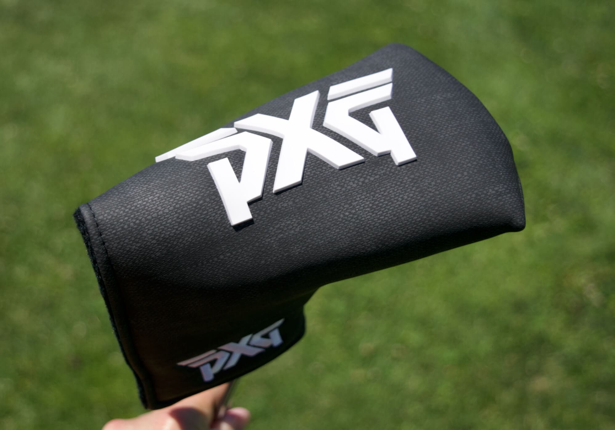芝生の上で手に持たれたPXGパター用ブラックヘッドカバー。大きなホワイトロゴが中央にデザインされている。