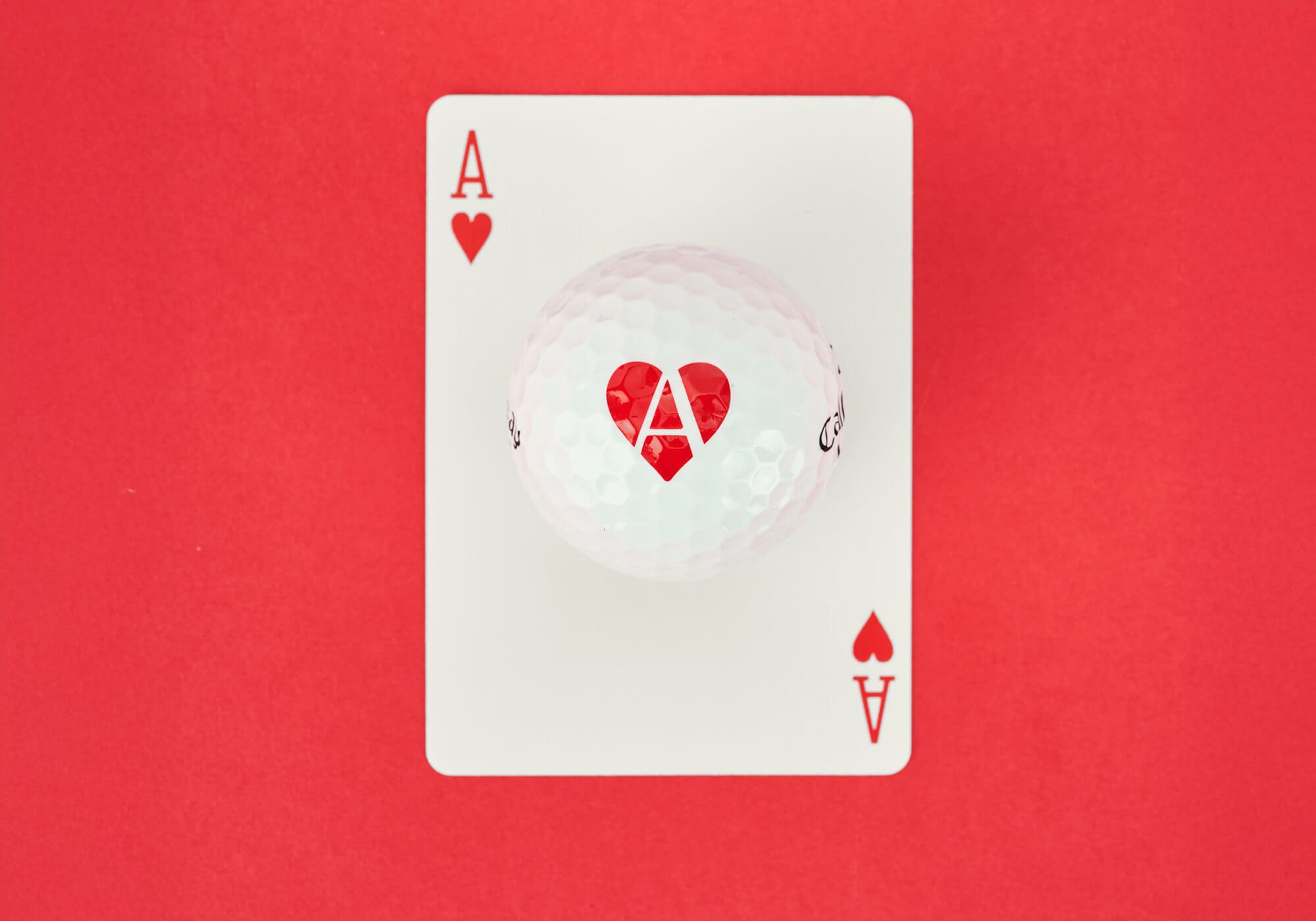 赤い背景に置かれたキャロウェイ「Supersoft ACES」ハート柄のゴルフボール。真下にはハートのエースカードが配置されている。