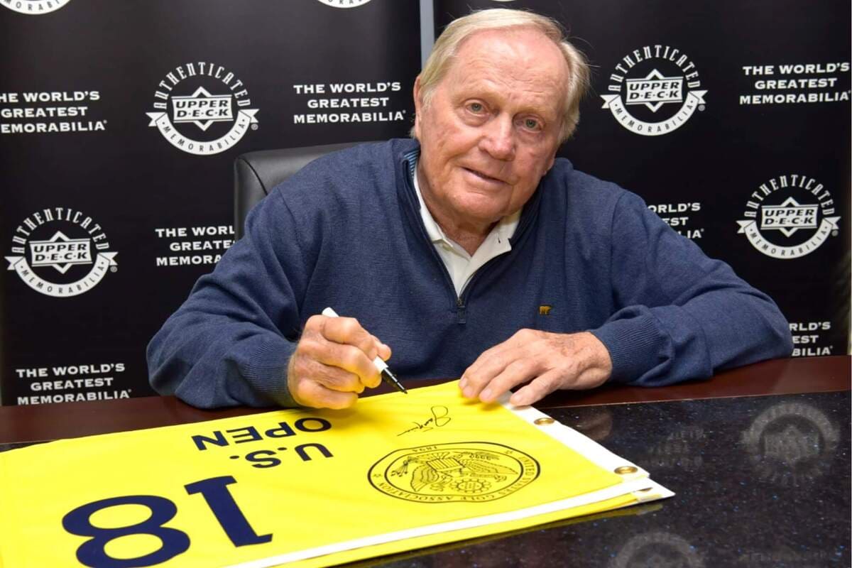 レジェンドゴルファーがサイン会で U.S. OPEN フラッグに直筆サインを書いている様子。背景にメモラビリアブランドのロゴ。