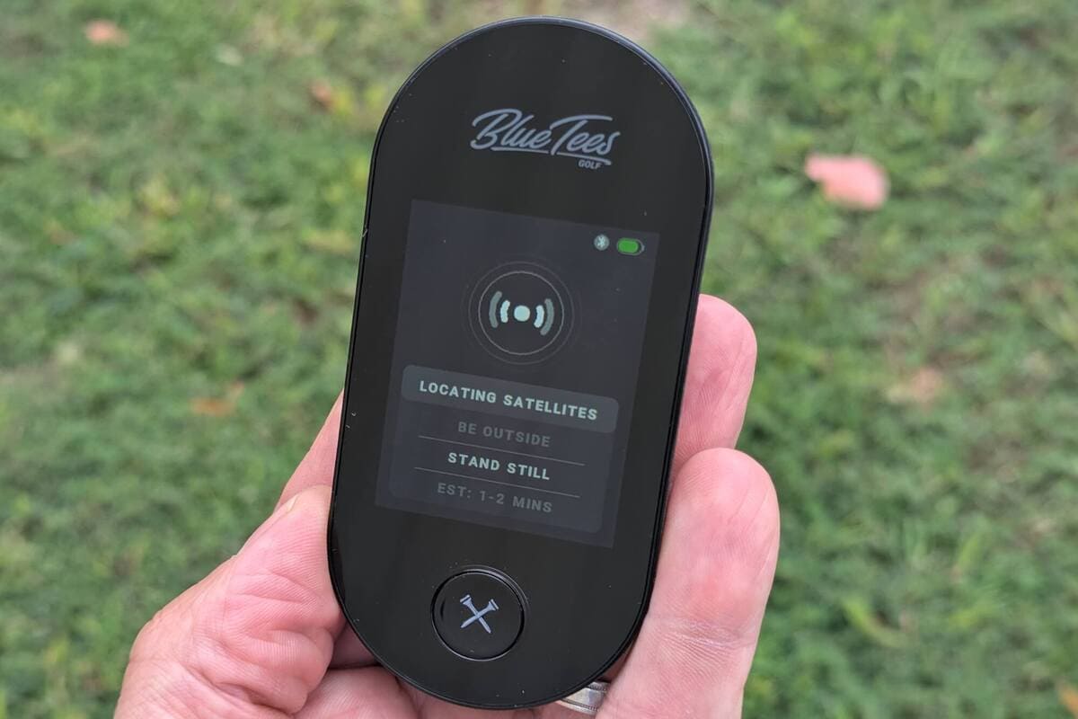 Blue Tees GolfのGPSゴルフナビが衛星を検索している様子。屋外での使用時に「LOCATING SATELLITES」と表示された画面を手に持っている状態。