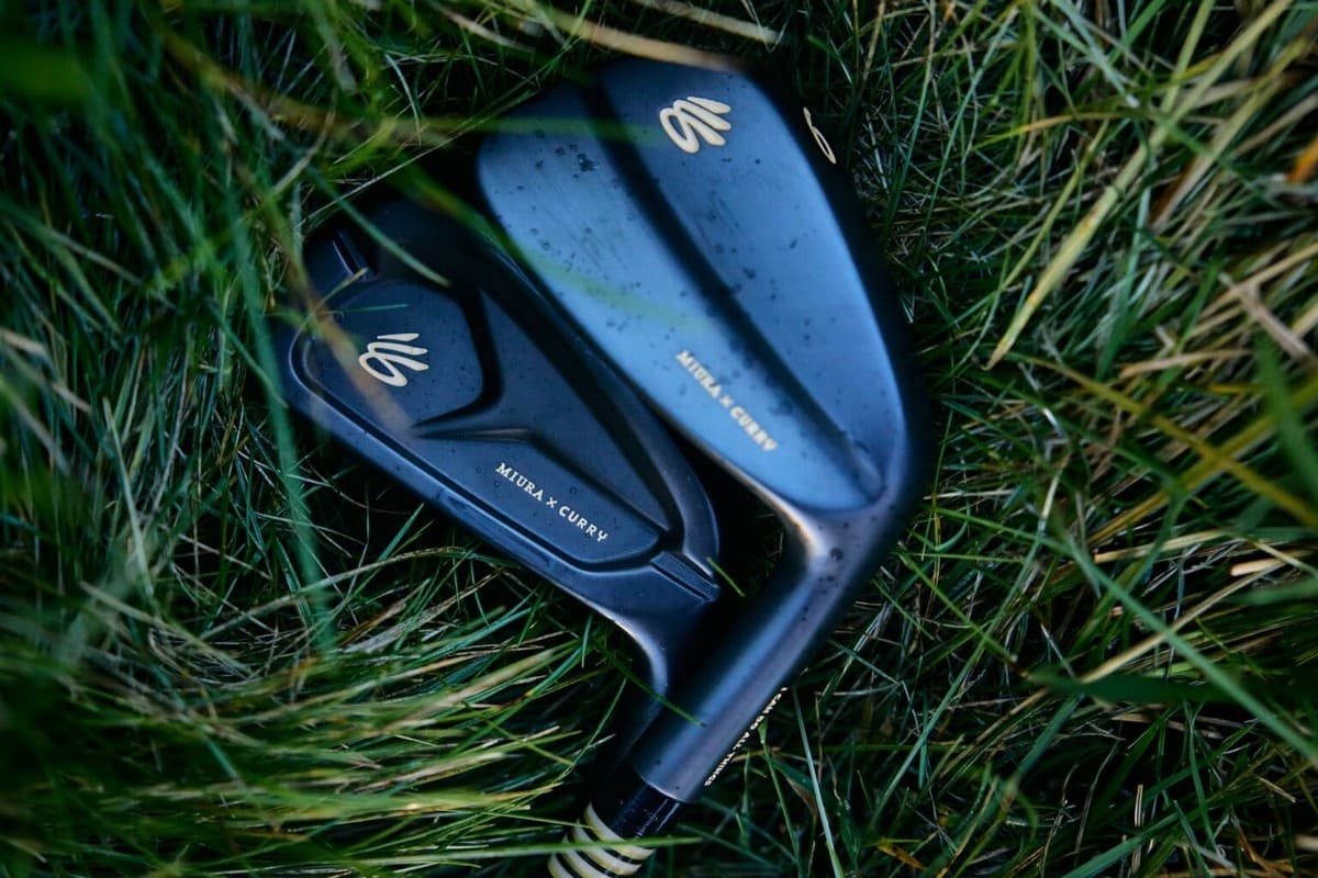 長い芝生の上に重ねて置かれたMiura x Curry限定アイアン2本。ダークカラーのキャビティバックとマッスルバックのヘッドに、金色のロゴと「MIURA × CURRY」の文字が見える。